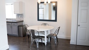 Een eethoek met een ronde tafel en vier stoelen, een keuken met kasten en een koelkast, en een deur aan de rechterkant.