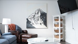 Een moderne woonkamer met een bank, een glazen salontafel, een berglandschap als wanddecoratie, een witte wandplank met manden en een flatscreen televisie.