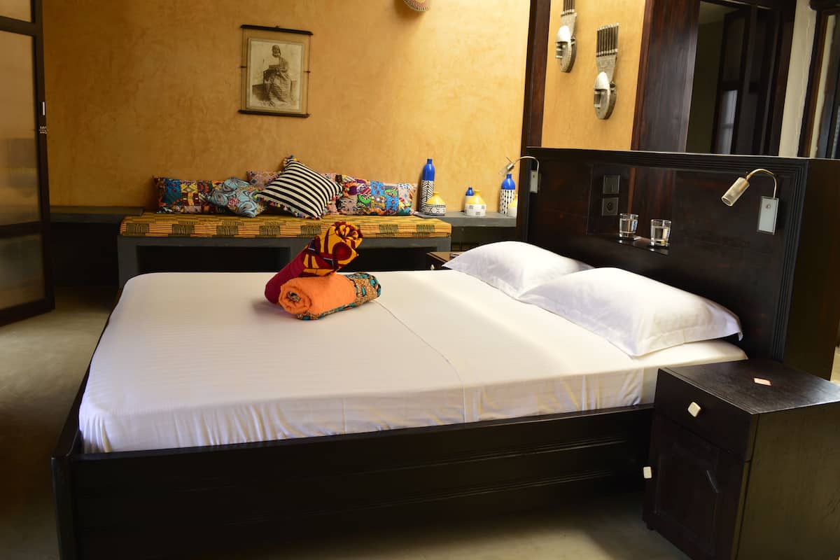 A hotel room with a bed, a bench with colorful cushions, a mirror, and a nightstand with a lamp.