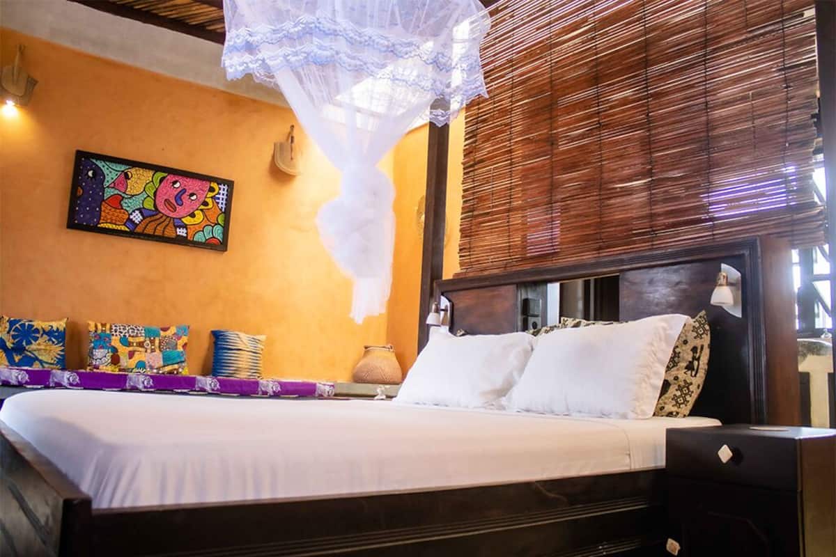 A hotel room with a bed, a colorful artwork on the wall, a mosquito net, and a window with blinds.