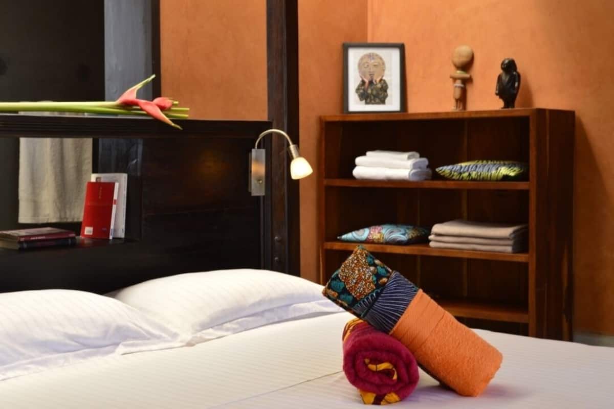 A neatly made bed with white linens, a bedside lamp, a wooden shelf with folded towels and decorative pillows, and a framed artwork on the wall.