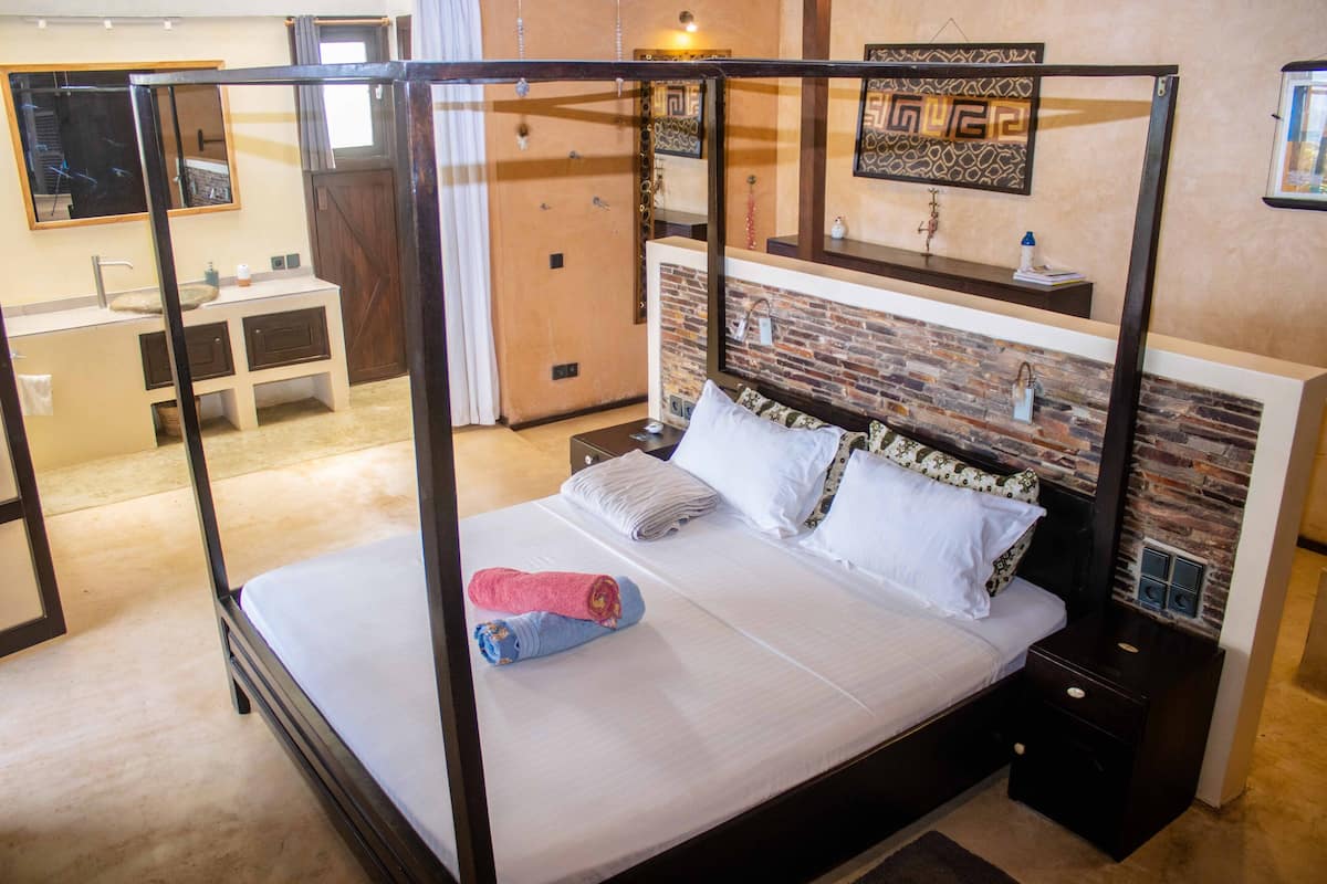 A four-poster bed with a headboard featuring a brick pattern, a nightstand with a lamp, and a bathroom with a sink and mirror.