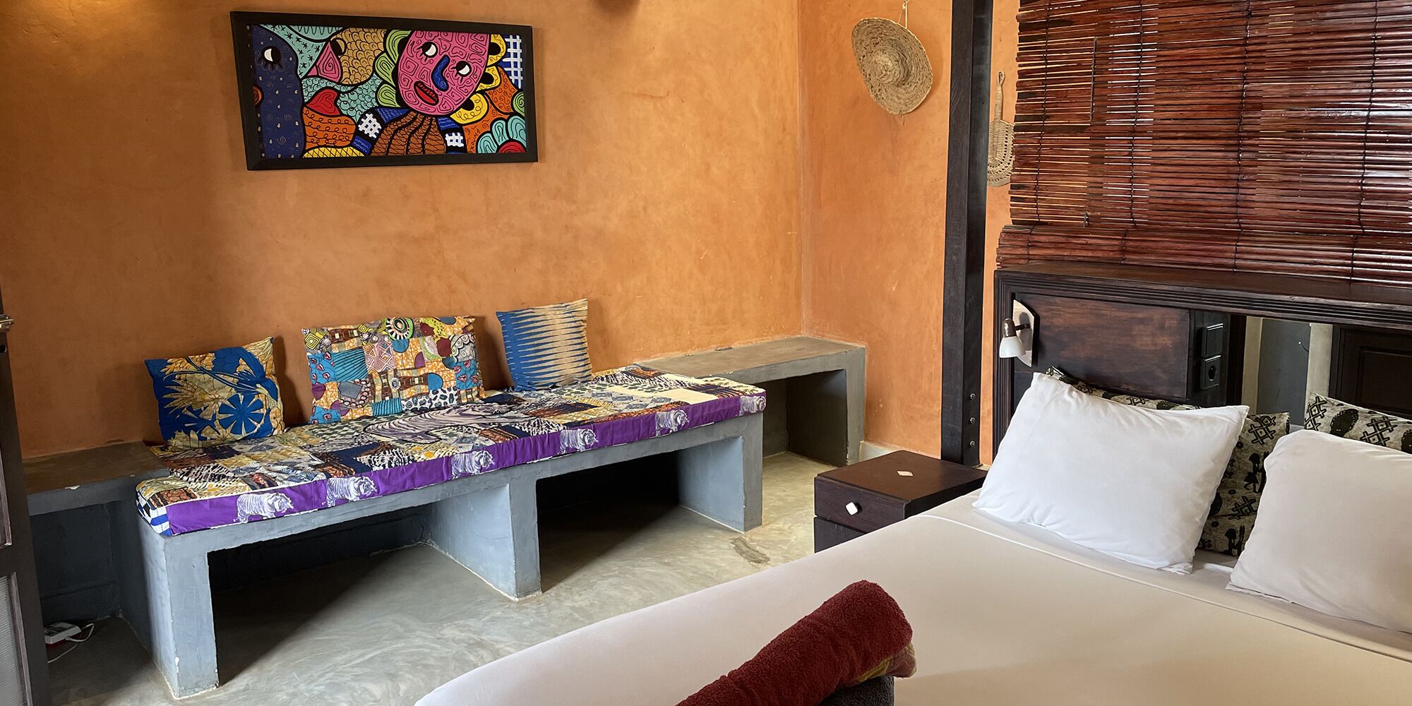 A hotel room with a bed, a bench with colorful cushions, a wall-mounted artwork, and a window with blinds.