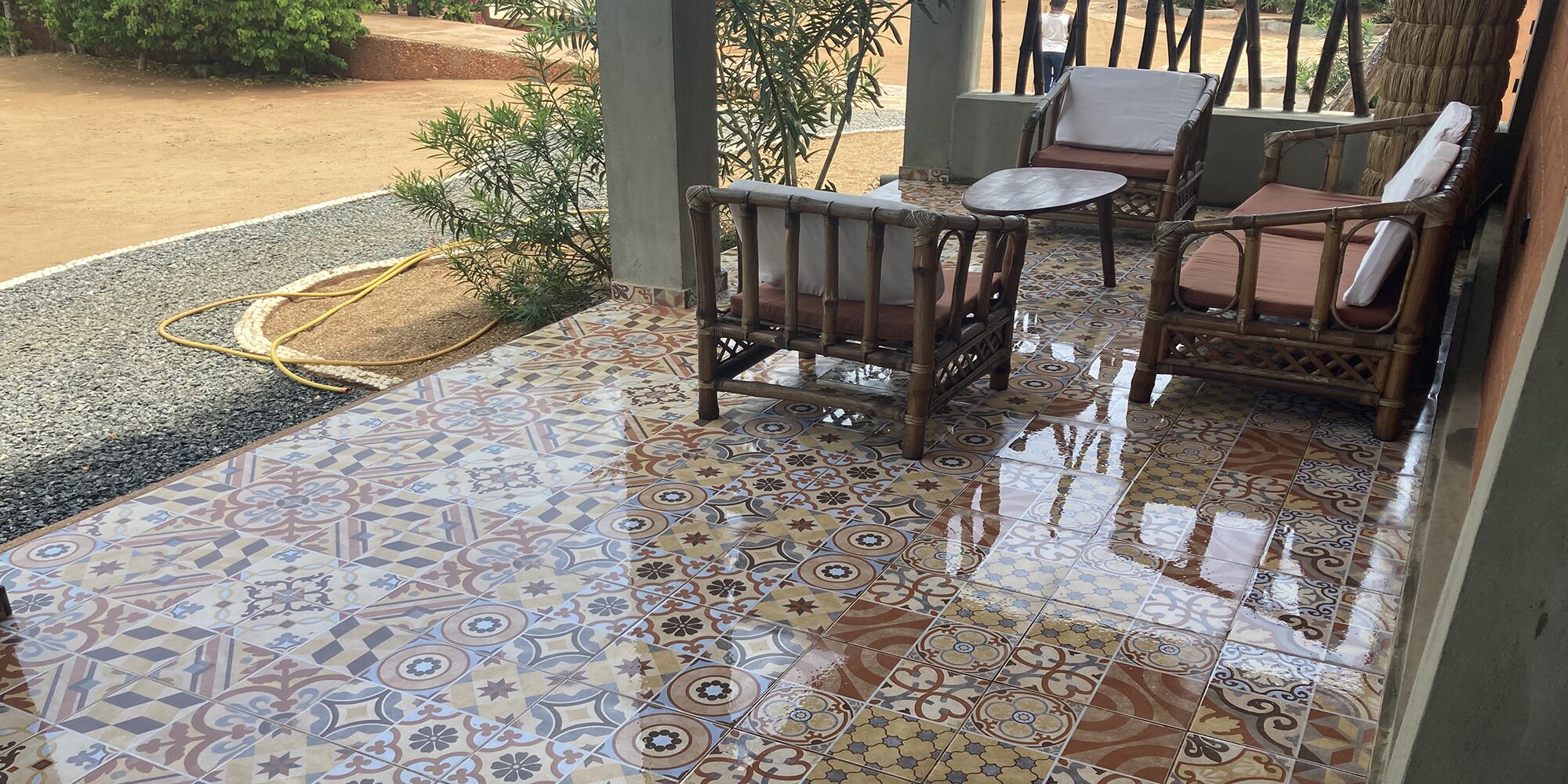 Un patio techado con baldosas estampadas, muebles de mimbre y una manguera en el suelo.