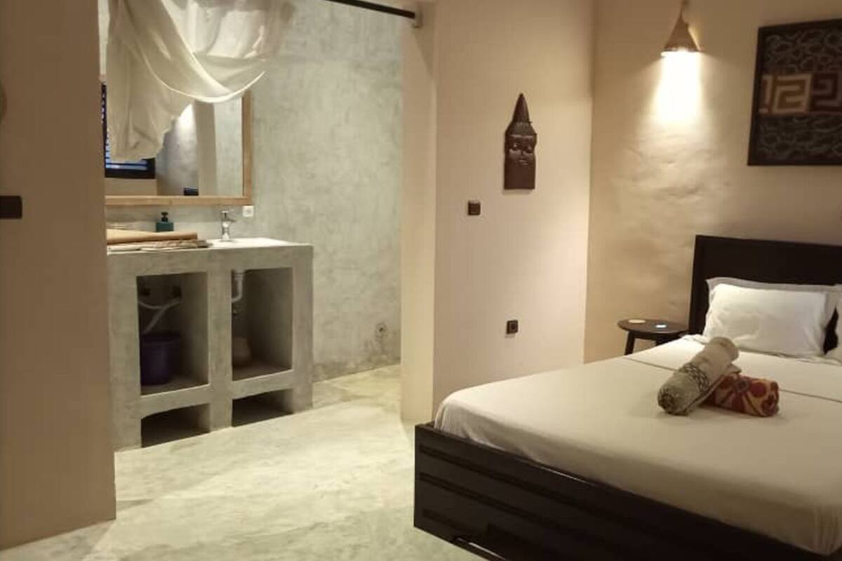 A hotel room with a bed, bedside tables, a nightstand, a mirror, a wall-mounted artwork, and a bathroom with a sink and cabinets.
