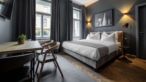 A modern bedroom with a bed, a desk, and a chair. There are large windows with curtains, a wall-mounted TV, and a floor lamp. - The Viaduct Suites & More (Prague)