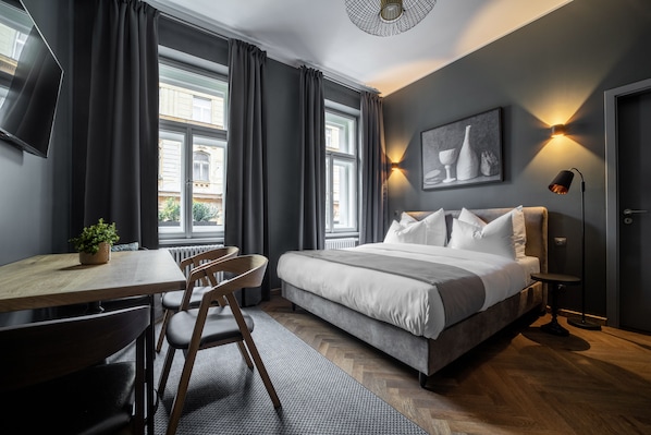 Quarto moderno com cama, escrivaninha e cadeira. Há janelas amplas com cortinas, uma TV fixada na parede e um abajur.