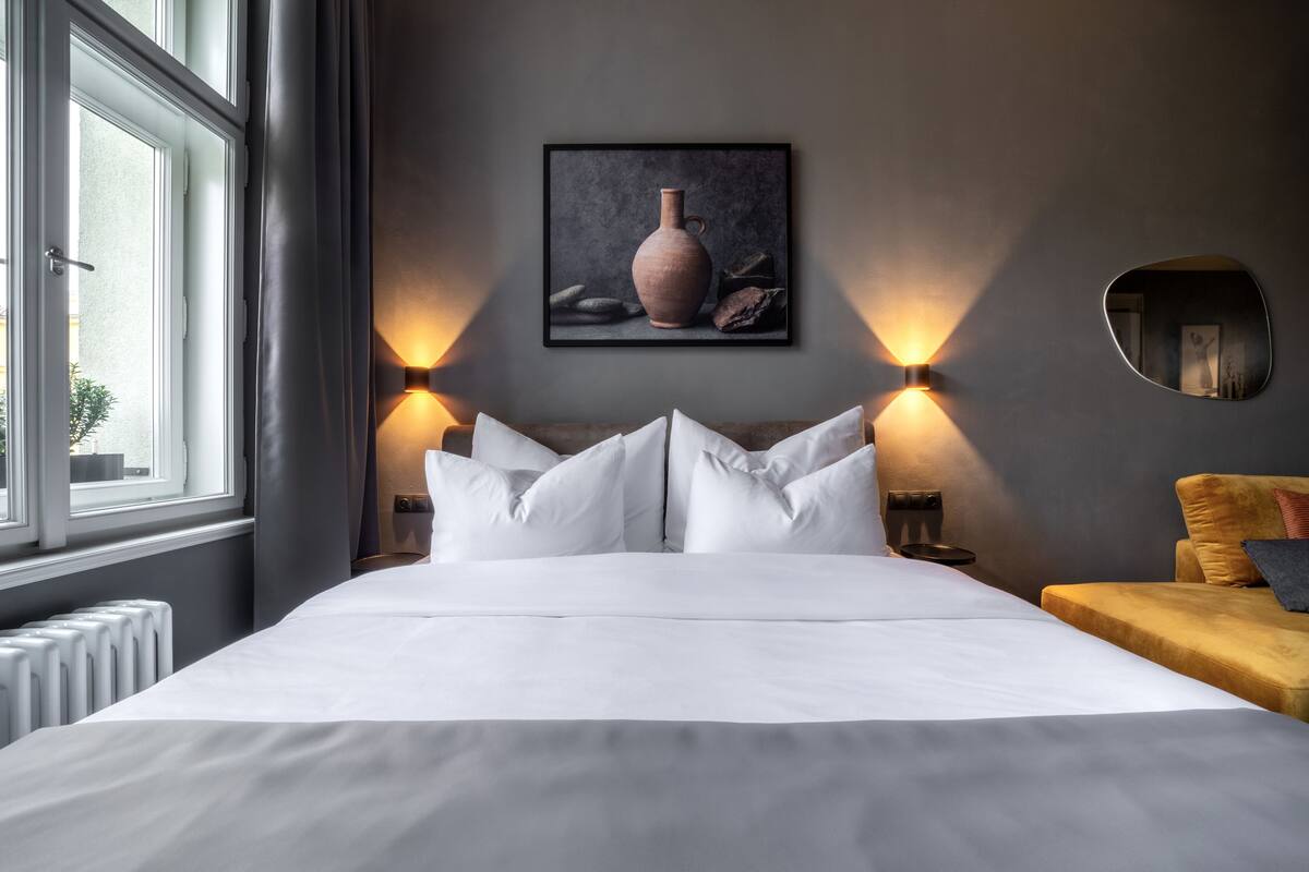 A hotel room with a large bed, two wall-mounted lamps, a framed artwork above the bed, and a window with curtains.
