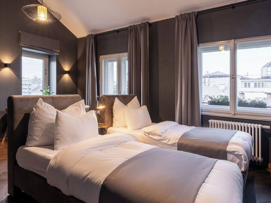 Una habitación de hotel moderna con una cama grande, dos mesitas de noche, un ventanal con cortinas y vistas a la ciudad.