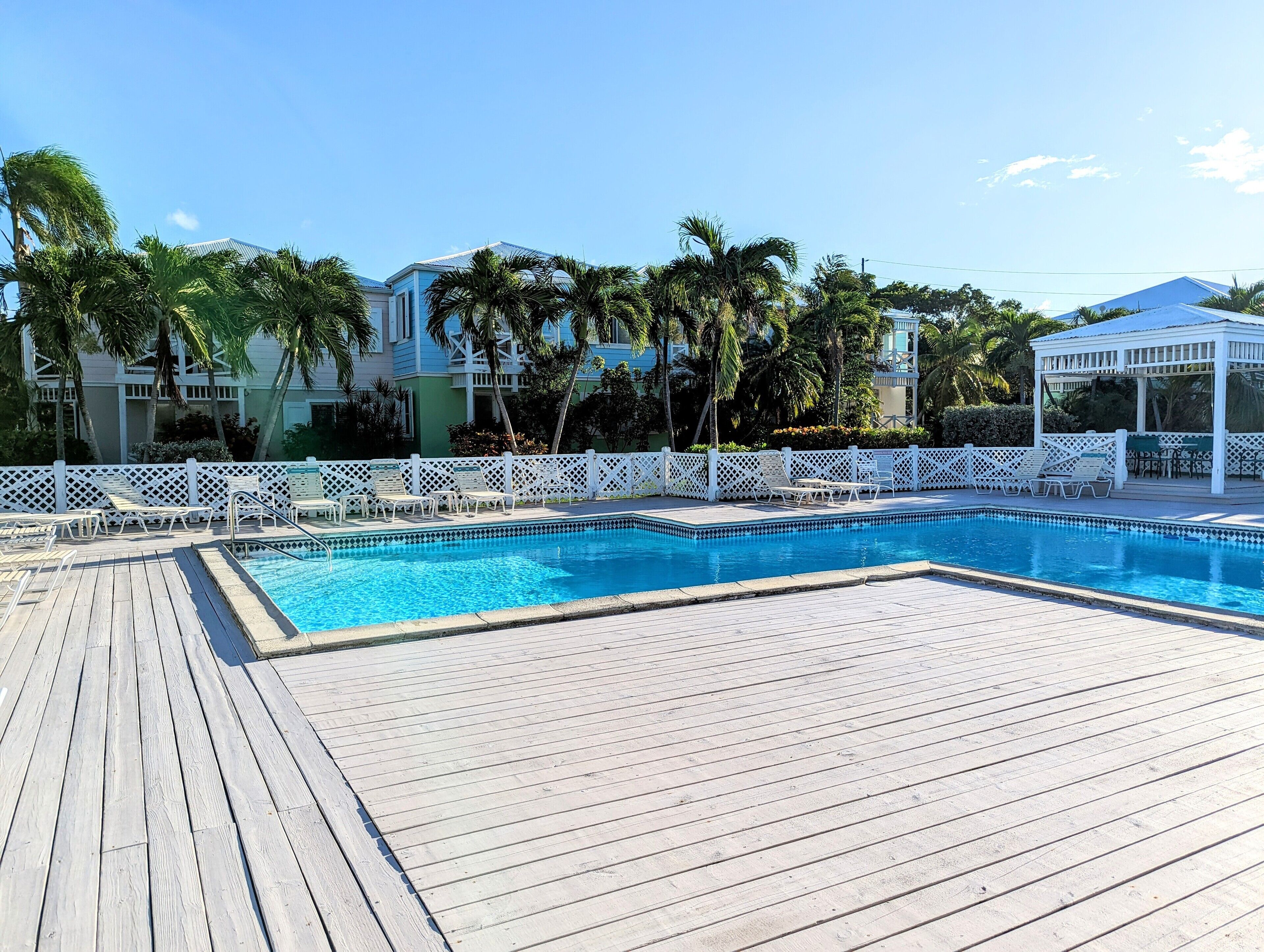 Zona de piscina con terraza de madera, valla de celosía blanca y sillones de descanso.
