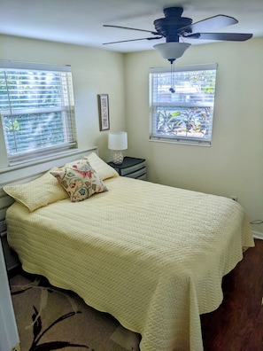 A bedroom with a bed, a ceiling fan, two windows with blinds, a bedside table with a lamp, and a framed picture on the wall.