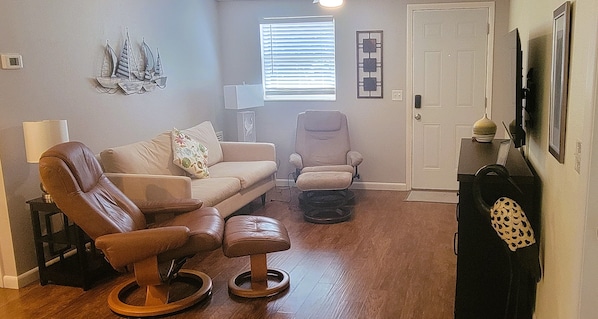 A living room with a brown leather sofa, a beige armchair, and a wooden ottoman. There is a door with a decorative mirror and a framed picture on the wall.