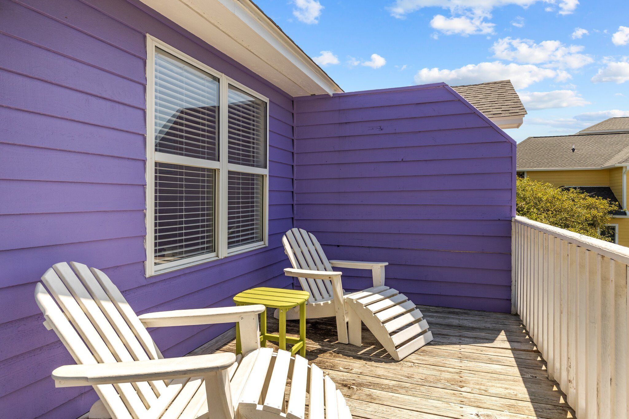Ein Balkon mit violetten Verkleidungen, zwei Adirondack-Stühlen und einem kleinen Tisch.