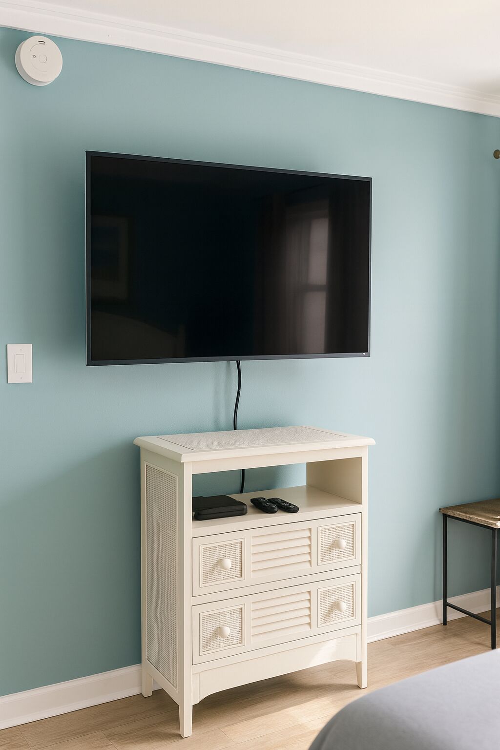 A room with teal walls, a mounted flat-screen TV, a white storage unit with drawers, and a small table.