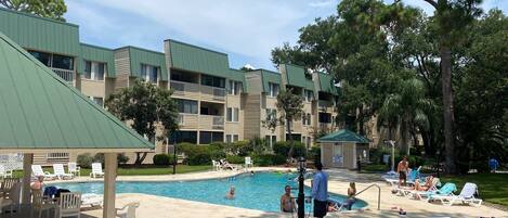 Zona de piscina con sillas de descanso y un área de estar cubierta, rodeada de edificios de varios pisos con balcones.
