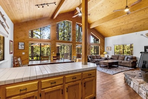 A spacious living area with a large wooden ceiling, a kitchen with a tiled countertop, and a stone fireplace. - Solitude in the Cedars, Spacious Mountain Home (Shaver Lake)