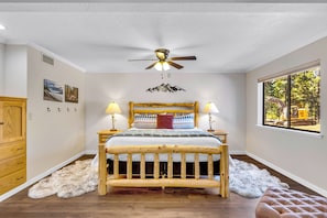 A bedroom with a wooden bed, two bedside lamps, a ceiling fan, and a window with a view of trees and a vehicle. - Solitude in the Cedars, Spacious Mountain Home (Shaver Lake)