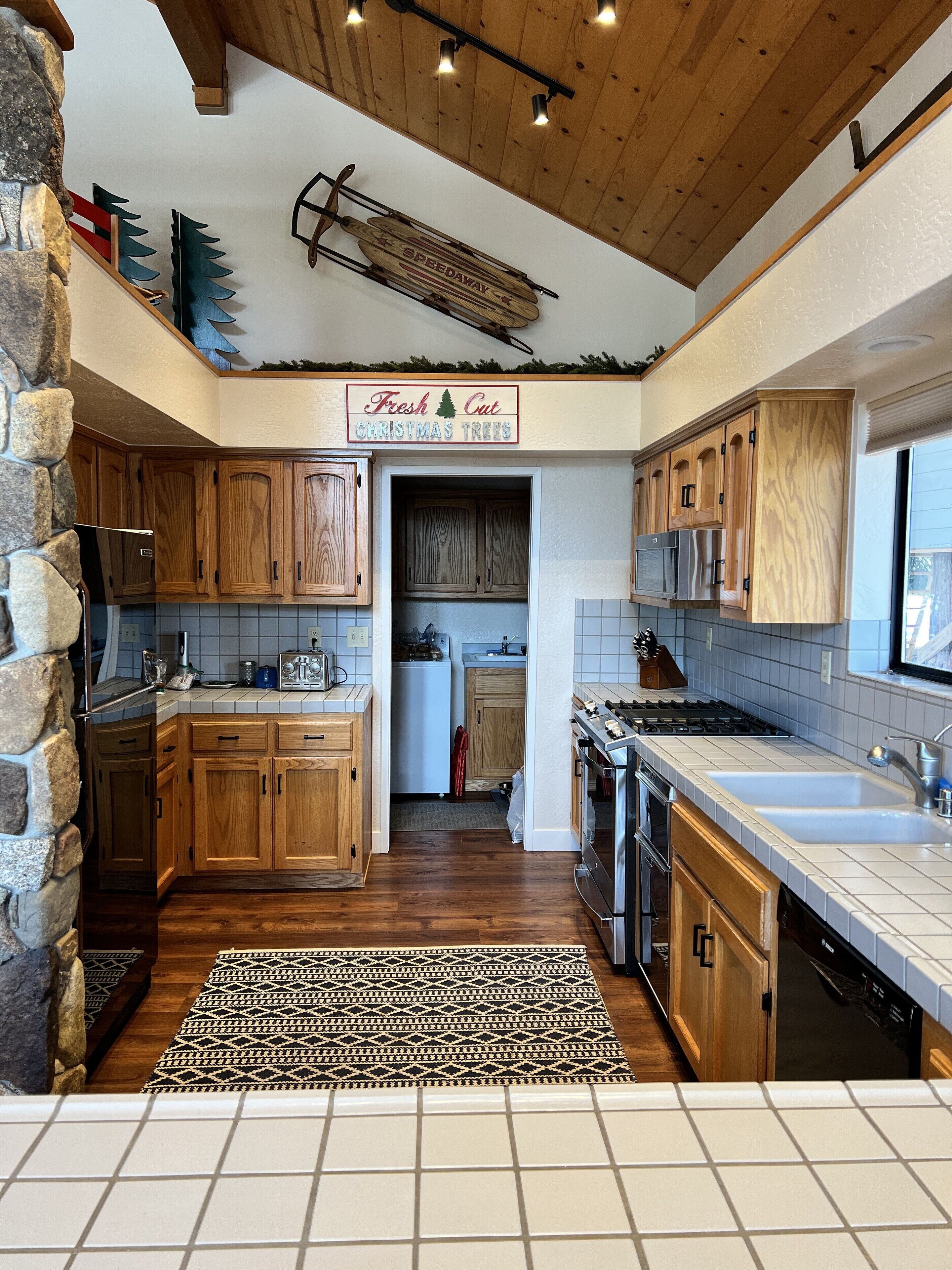 A kitchen with wooden cabinets, a stone accent wall, a patterned rug, and a sign indicating fresh cut Christmas trees.