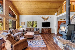 A spacious living room with a large wooden ceiling, a fireplace, a television, and a stone fireplace surround. - Solitude in the Cedars, Spacious Mountain Home (Shaver Lake)