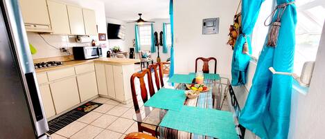 A compact kitchen with a microwave, stove, and cabinets. A dining table with chairs is set with a fruit bowl and placemats.