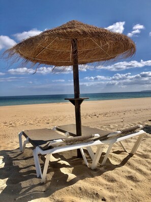 A beach with a straw umbrella and white lounge chairs.