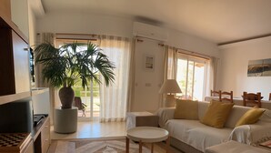 Uma sala de estar com sofá, mesa de centro, televisão, planta e janela com cortinas.