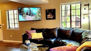 A living room with a black leather sofa, a glass coffee table, and a colorful rug. There is a ceiling fan, a TV mounted on the wall, and a window with a view of trees. - Lovely Downtown Condo, Walk to Historic Downtown! (Flagstaff)