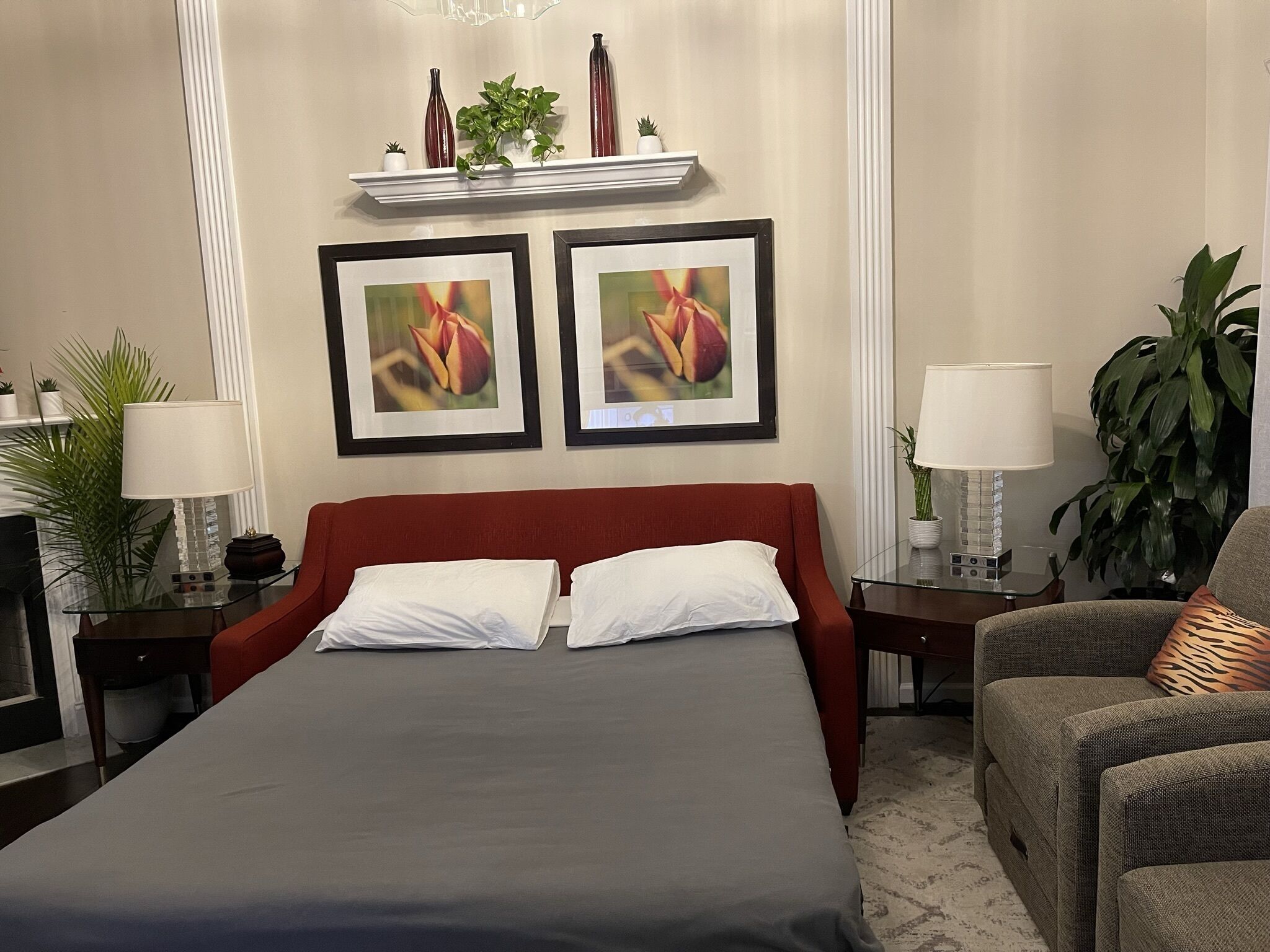 Habitación de hotel con una cama grande, dos cuadros enmarcados con motivos florales, cabecera roja, una mesa de centro de vidrio, una mesita de noche con lámpara, una silla y una planta en maceta.