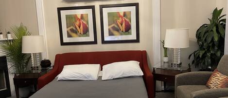 A hotel room with a large bed, two framed floral paintings, a red headboard, a glass coffee table, a side table with a lamp, a chair, and a potted plant.