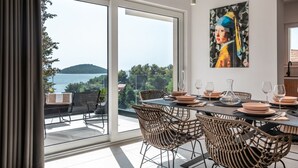 Un coin repas avec une table dressée pour six personnes, de grandes fenêtres offrant une vue sur la mer et les collines, et une œuvre d’art moderne accrochée au mur.