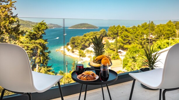 Un balcon avec deux chaises blanches, une petite table sur laquelle se trouve une assiette de croissants, un verre de vin rouge et un saladier rempli d’ananas, de bananes et d’oranges.