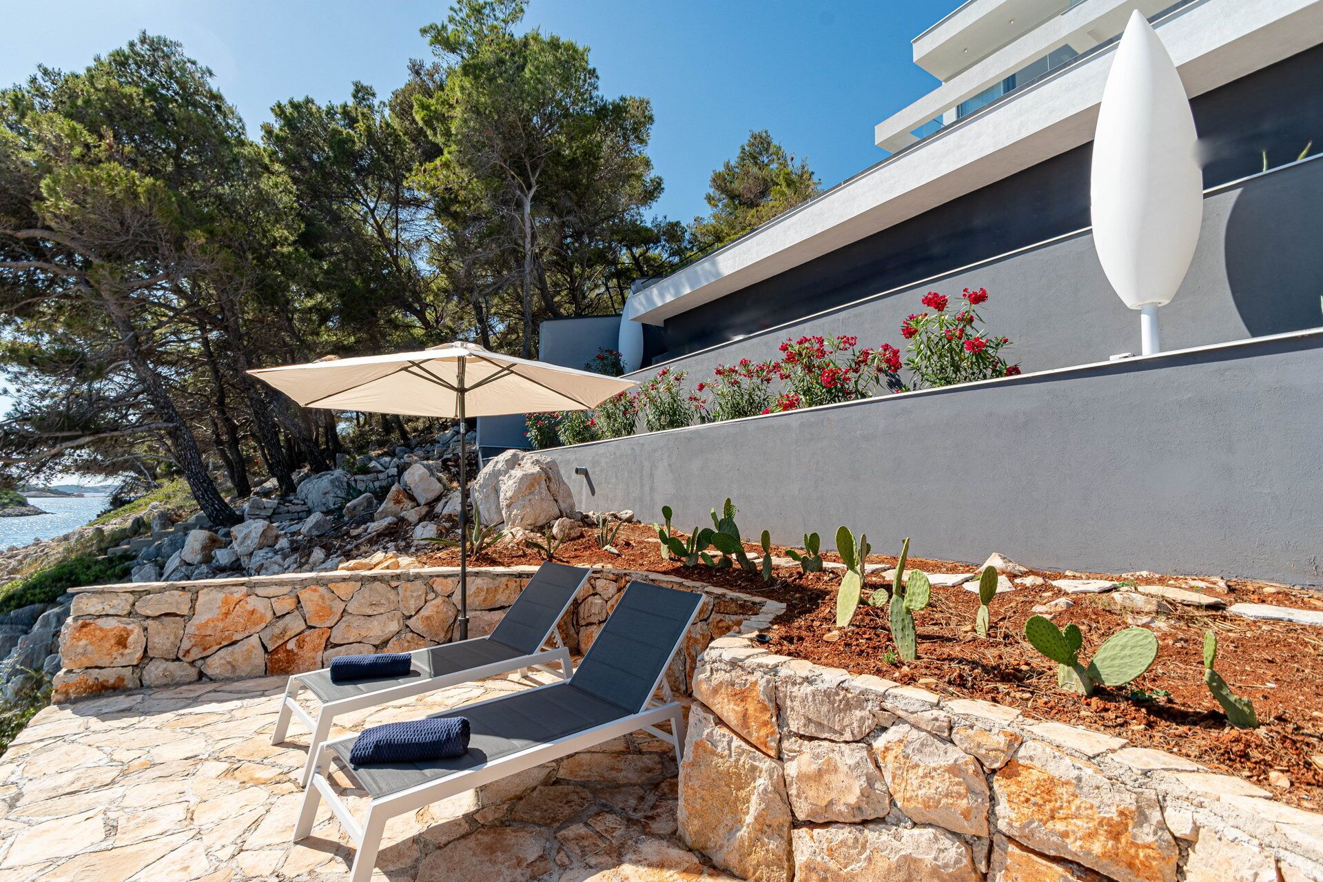 Un patio de piedra con dos sillones de descanso y una sombrilla color beige, con vista a una costa rocosa y un edificio moderno con pared blanca y flores rojas.