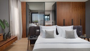 Quarto moderno com cama grande, mesa de cabeceira, espelho e vista para o banheiro por uma porta de vidro.