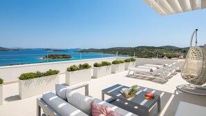 Une terrasse sur le toit, meublée de mobilier blanc, avec une table sur laquelle sont disposés un plateau de livres et un vase, et d’où l’on peut admirer la mer et les collines.