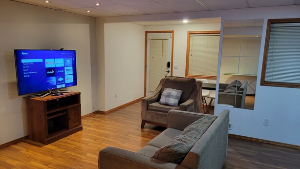 Una sala de estar con una televisión de pantalla plana sobre un soporte de madera, un sofá gris y un sillón marrón.