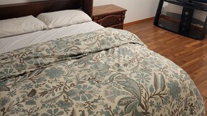 Un dormitorio con cama, una mesita de noche de madera, un televisor mostrando 'NETFLIX' y una ventana con persianas.