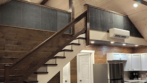 A spacious loft with a wooden staircase, a plush brown armchair, a stainless steel refrigerator, and a kitchen area.