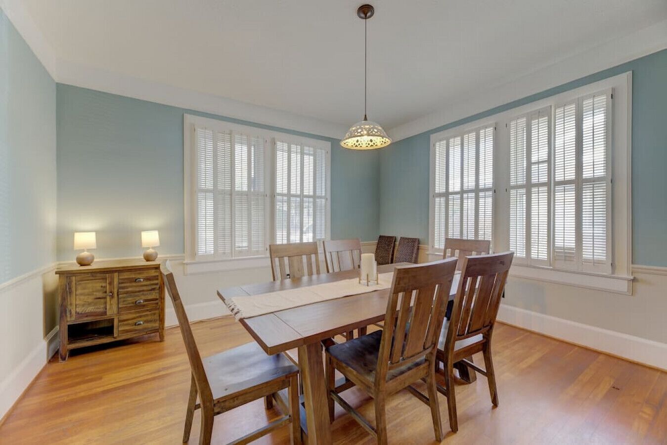 A dining area with a wooden table and chairs, white window shutters, and a hanging light fixture.