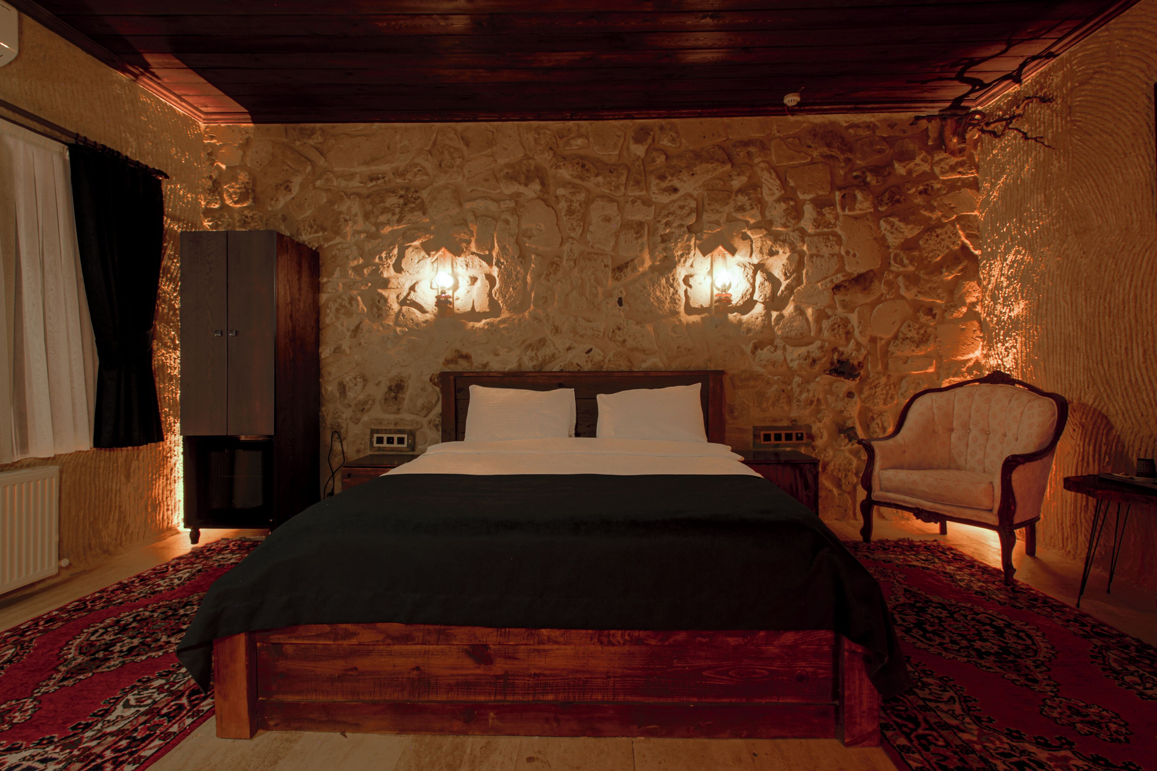 A bedroom with a stone wall, a wooden bed, a chair, and two wall-mounted lamps.