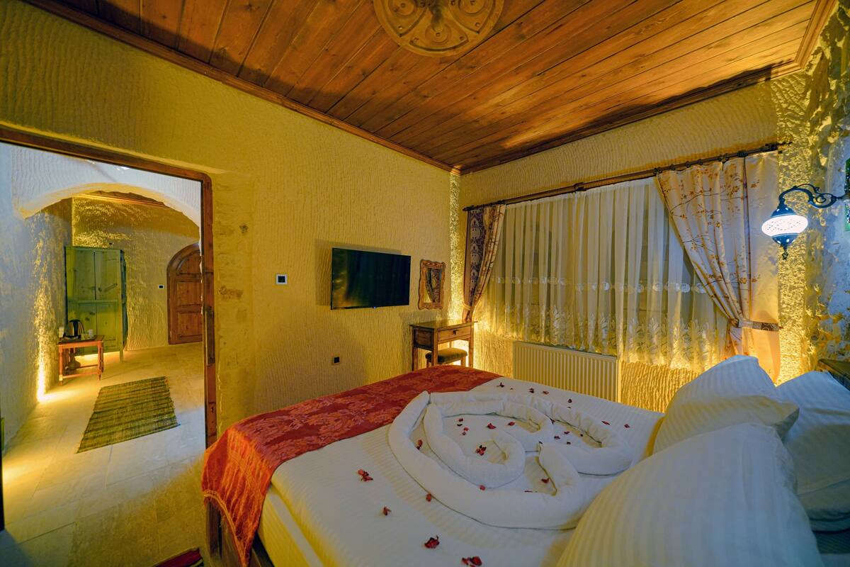 A cozy bedroom with a wooden ceiling, a bed with a red blanket, a television on the wall, and a window with curtains.