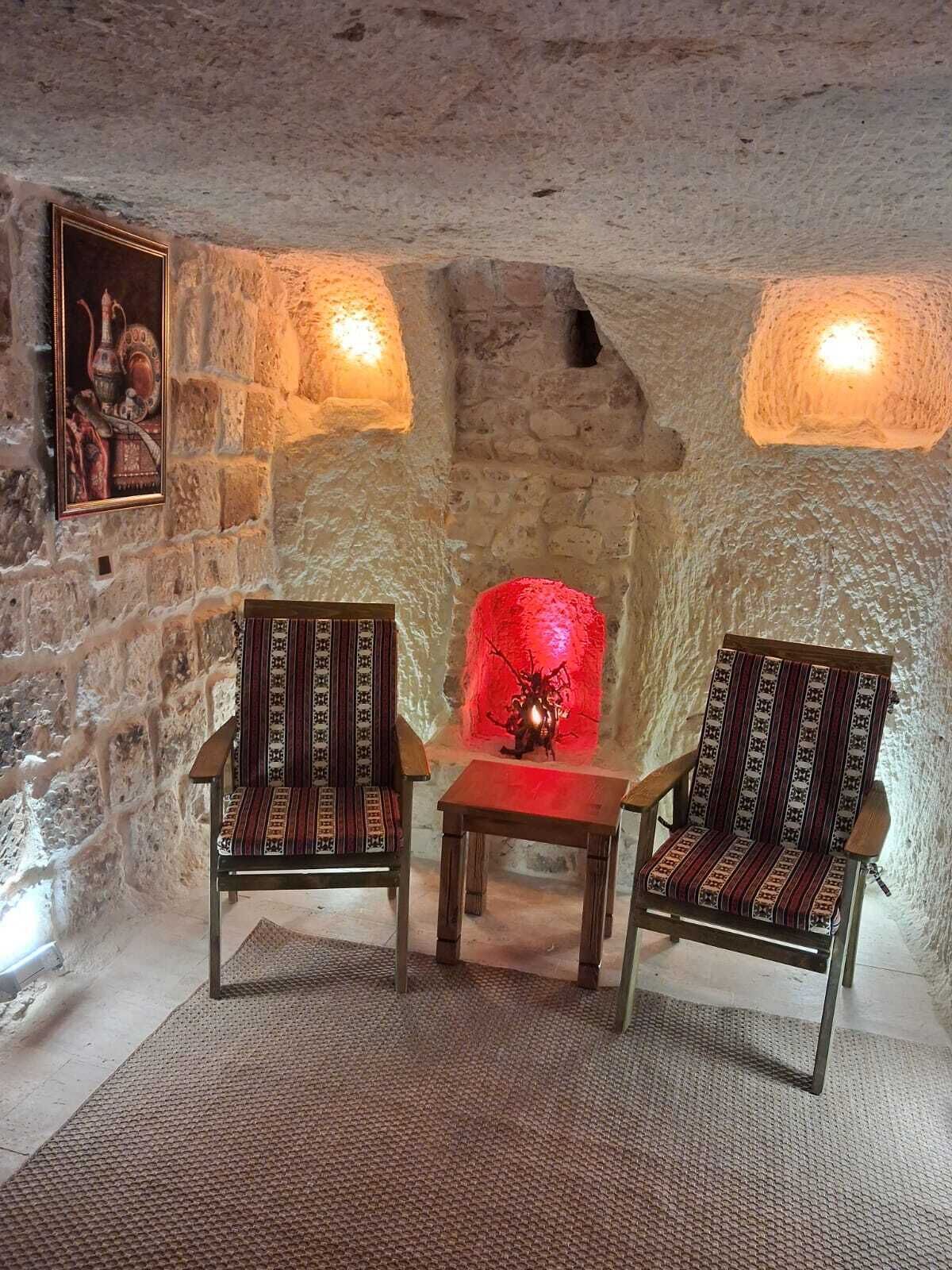 Ein Zimmer mit Steinwänden, zwei Holzstühlen und einem Tisch, einem gerahmten Bild an der Wand und einem roten Dekorationsobjekt in der Mitte.