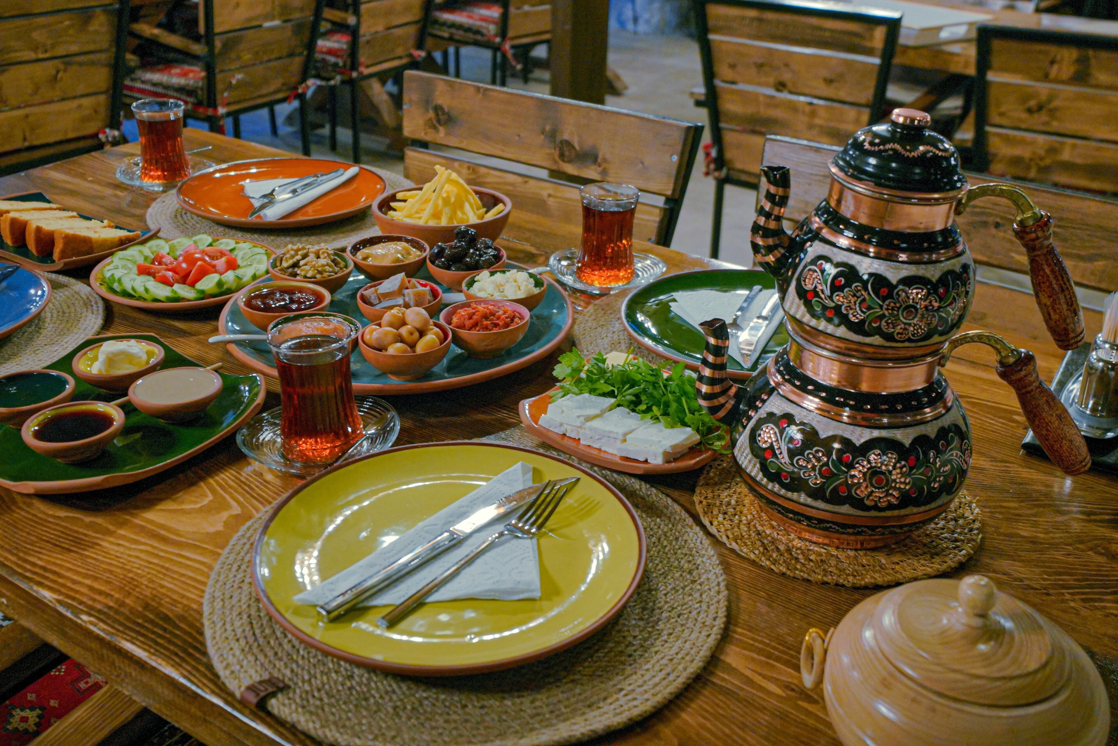 A wooden dining table set with various dishes, including salads, bread, and a traditional copper teapot.