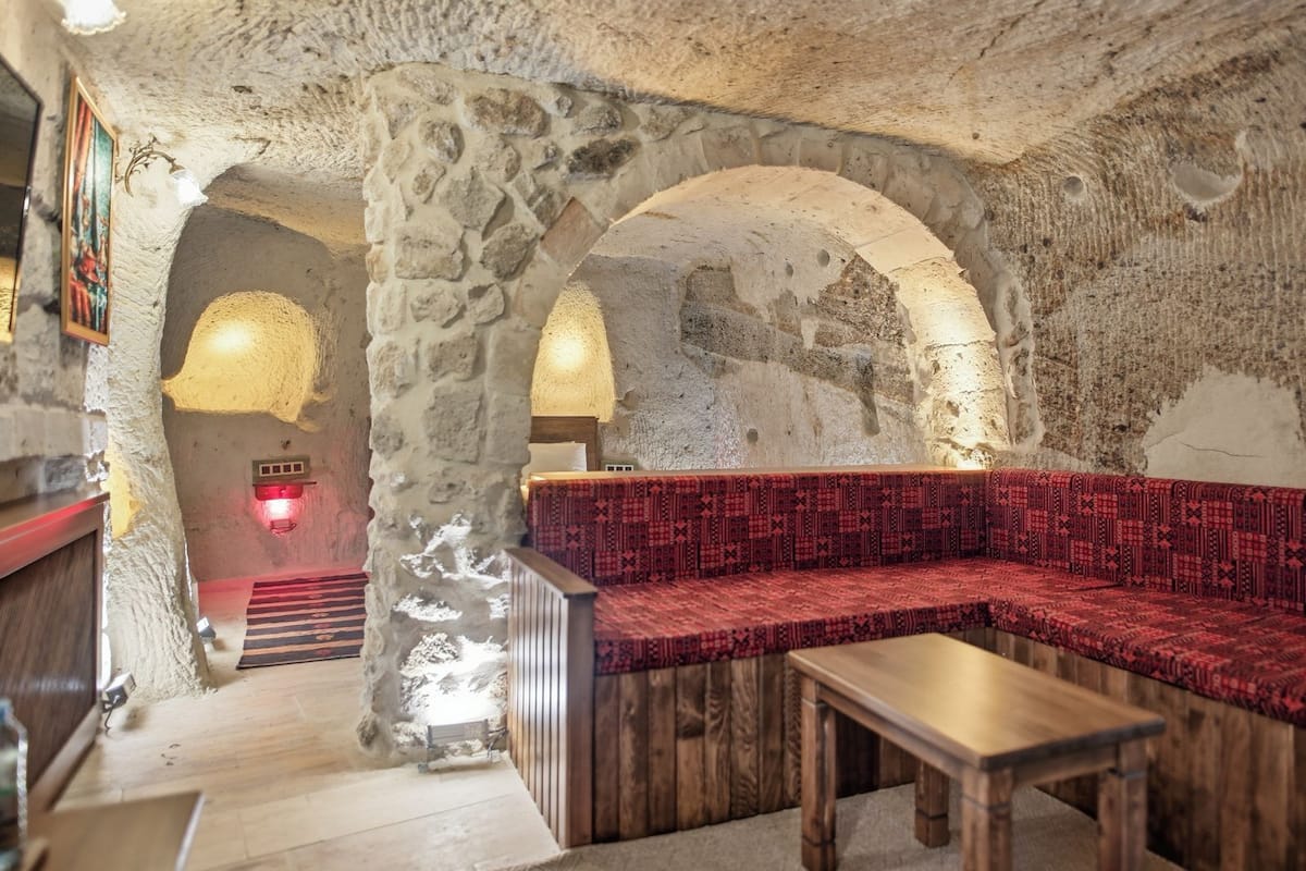 A stone-walled interior with a wooden bench and table, a patterned red sofa, and a red light fixture.