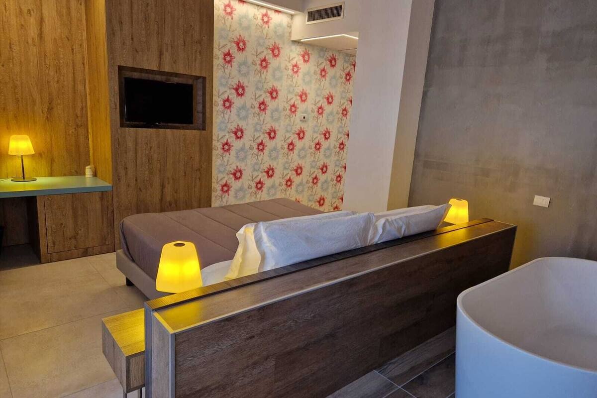 A modern bedroom with a wooden headboard, a flat-screen TV mounted on the wall, a floral-patterned wallpaper, and a large bathtub.