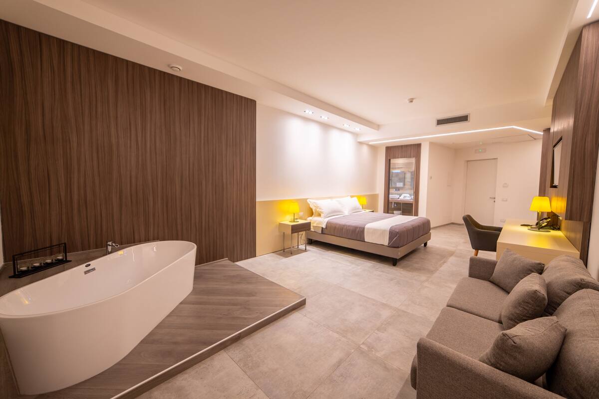A modern hotel room with a large bathtub, a bed, a sofa, and a desk with a lamp.