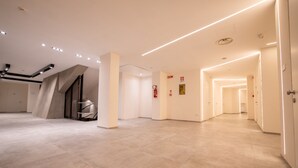 Un vestíbulo de hotel amplio y moderno, con un diseño limpio y minimalista, que presenta una escalera central, un largo pasillo con puertas y un suelo de baldosas.