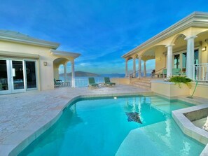 Una piscina con vista despejada al océano, rodeada por un porche cubierto con sillas y una escalera que lleva a un nivel superior.