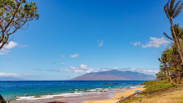 Ein Strand mit einer Holzterrasse, einem blauen Sonnenschirm und einem Berg im Hintergrund.