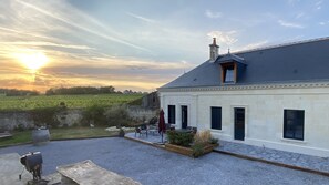 A building with a stone facade, a sloped roof, and a chimney. There is a gravel patio with outdoor furniture and a small garden area. - Cocoon of the vines, Maison Tourangelle in the heart of Domaine Ansodelles (Benais)