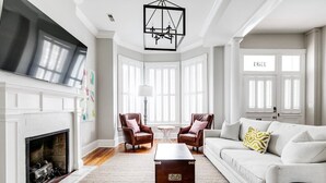 Een ruime woonkamer met een open haard, een grote flatscreen televisie, leren fauteuils, een witte bank en een kroonluchter.